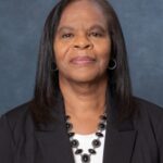 A woman with long, straight hair dons a black blazer over a white top, complemented by a black beaded necklace. She stands confidently against a plain blue-gray backdrop, embodying the sophistication of Brendolyn elegance.