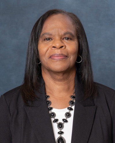 A woman with long, straight hair dons a black blazer over a white top, complemented by a black beaded necklace. She stands confidently against a plain blue-gray backdrop, embodying the sophistication of Brendolyn elegance.