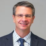 A man with short gray hair and glasses, wearing a dark suit, checkered shirt, and dotted tie, smiles and looks at the camera against a plain light gray background.