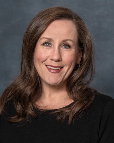 Katie Ellison, a woman with long brown hair, smiles at the camera. She is wearing a black top against a neutral background, exuding warmth and charm.