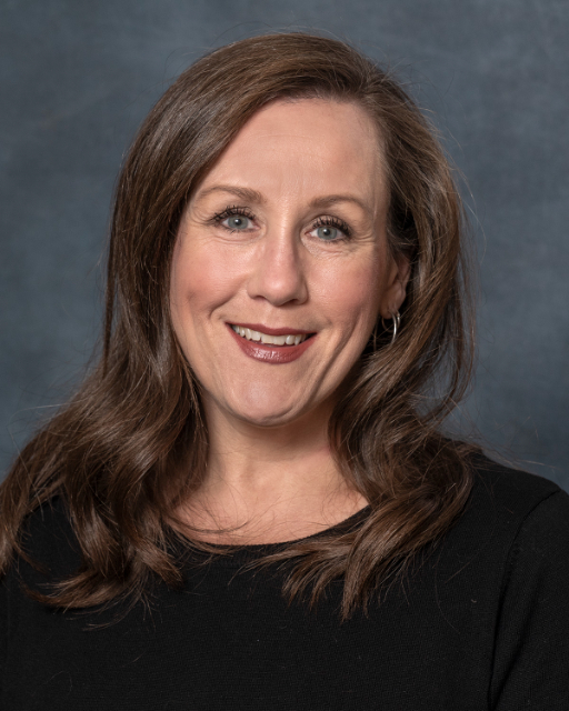 Portrait of a woman with long, wavy brown hair and hoop earrings, smiling at the camera. She is wearing a black top and is set against a textured gray background. Her name is Katie Ellison, embodying warmth and elegance in every detail.