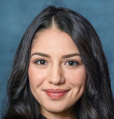 Aila, a woman with long dark hair and dimpled cheeks, smiles warmly at the camera against a blue background. Her natural makeup enhances her radiant features.