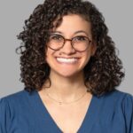 Brittany, with her curly hair and glasses, smiles warmly. She wears a blue V-neck top and a simple necklace, exuding friendliness against the plain gray background.