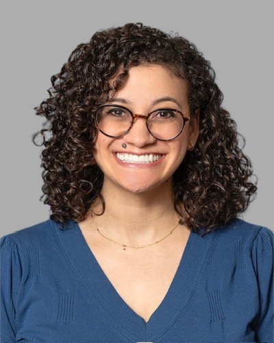 Brittany, with her curly hair and glasses, smiles warmly. She wears a blue V-neck top and a simple necklace, exuding friendliness against the plain gray background.