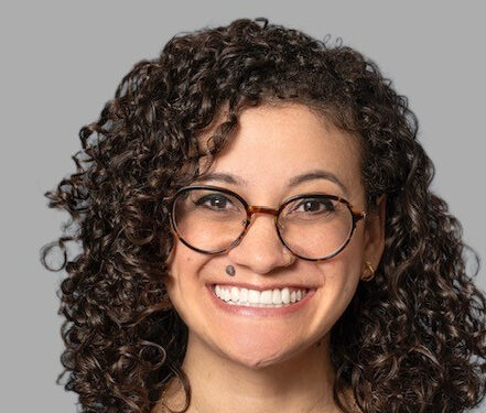 A smiling person with curly hair and round glasses is set against a plain gray background, exuding an effortless charm reminiscent of Brittanys vivacious style.