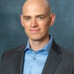 Gregory, with his head shaved, wears a blue shirt and dark blazer. His slight smile adds warmth to the dark gray backdrop.