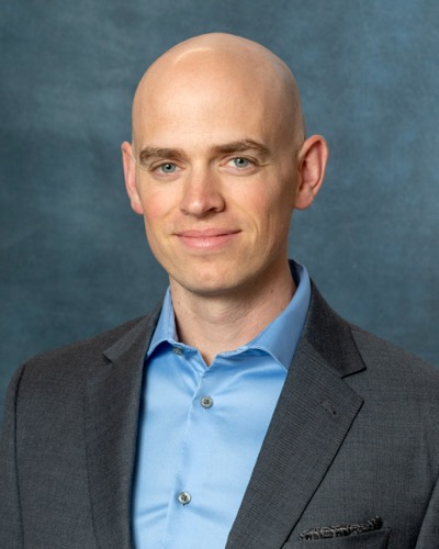 Gregory, with his head shaved, wears a blue shirt and dark blazer. His slight smile adds warmth to the dark gray backdrop.