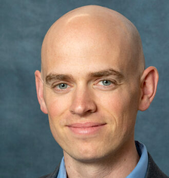 Gregory, a bald man in a suit with a light blue shirt, poses against a textured gray background, smiling slightly.
