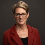 Jennifer, with short hair and glasses, wearing a red jacket over a black top, smiles warmly against the dark background. Her expression suggests shes just heard a delightful story from one of her favorite books.