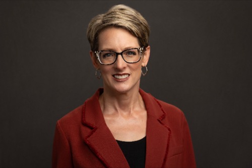 Jennifer, with short hair and glasses, wearing a red jacket over a black top, smiles warmly against the dark background. Her expression suggests shes just heard a delightful story from one of her favorite books.