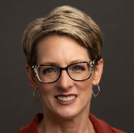 A person with short hair is wearing glasses and hoop earrings, smiling in front of a dark background, reminiscent of someone who spends hours enjoying books.
