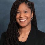 LaToshia Boyd-Lee, with long curly hair, smiles while wearing a black blazer and a necklace with a cross pendant. The background is a muted blue-gray.