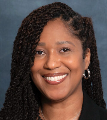 Portrait of LaToshia, a smiling person with long, braided hair and hoop earrings, against a plain, dark blue background. They are wearing a dark top.