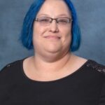 Susan, with her vibrant blue hair and glasses, wears a black top as she smiles warmly at the camera against a gray background.