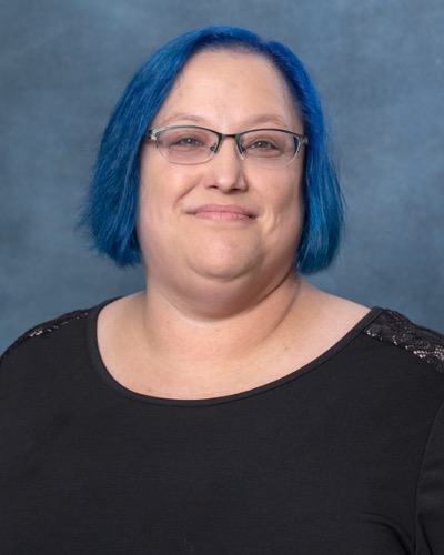 Susan, with her vibrant blue hair and glasses, wears a black top as she smiles warmly at the camera against a gray background.