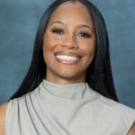 Morgan, a woman with long straight black hair, smiles against a blue background. She is wearing a gray sleeveless top with a high neckline.