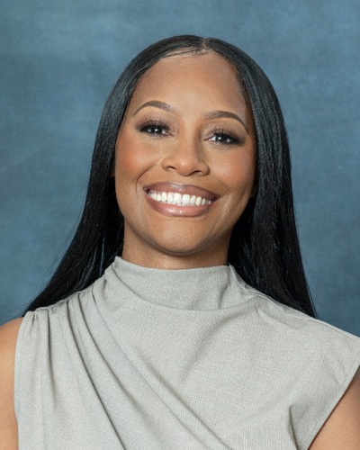 Morgan, a woman with long straight black hair, smiles against a blue background. She is wearing a gray sleeveless top with a high neckline.