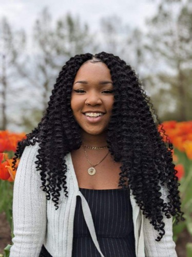 A person with long curly hair smiles while standing in a field of vibrant orange flowers, reminiscent of Dajes serene landscapes. They wear a white cardigan over a dark dress and a necklace with a pendant. The background shows blurred greenery and flower stems.