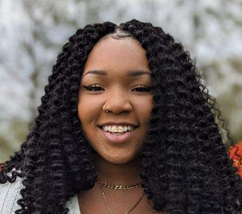 A person with long curly hair smiles at the camera, exuding a Bradshaw-Glenn charm. They are wearing a white top and a gold necklace. The background is blurred, hinting at greenery and light sky.