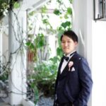 A person in a formal navy suit, white shirt, and bow tie stands casually against a wall in a well-lit corridor with greenery. They have a boutonniere on their lapel and are gazing towards the camera.