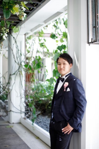 A person in a formal navy suit, white shirt, and bow tie stands casually against a wall in a well-lit corridor with greenery. They have a boutonniere on their lapel and are gazing towards the camera.