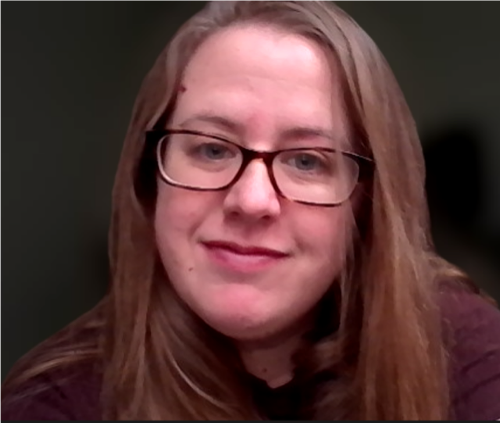 Bridget, with her long brown hair and glasses, is smiling softly against a dark background. She is wearing a brown top and appears relaxed, exuding an aura of quiet confidence typical of the Coffey family.