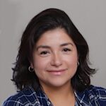 Paola, with shoulder-length dark hair, is smiling warmly in her blue and white plaid shirt. The background is plain and neutral, emphasizing her cheerful demeanor.