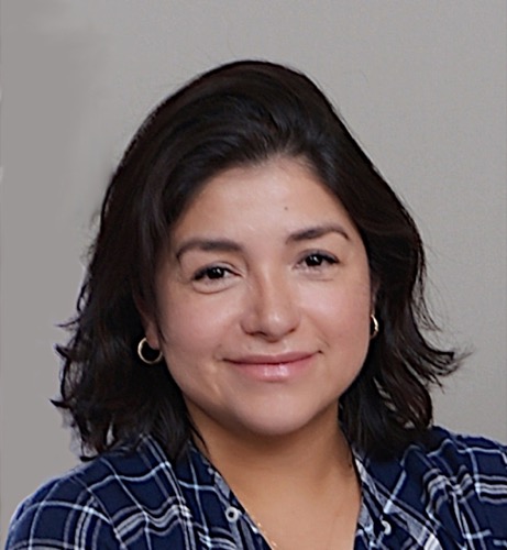 Paola, with shoulder-length dark hair, is smiling warmly in her blue and white plaid shirt. The background is plain and neutral, emphasizing her cheerful demeanor.