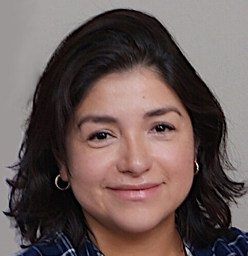 Paola, with short, dark hair and hoop earrings, smiles warmly at the camera. Clad in a dark checkered shirt, she stands against a plain light-colored background, exuding effortless charm.