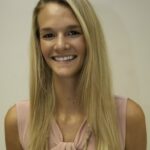 Lauren, with her long, straight blonde hair and sleeveless beige top, smiles at the camera against a plain, light-colored wall.