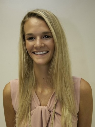 Lauren, with her long, straight blonde hair and sleeveless beige top, smiles at the camera against a plain, light-colored wall.