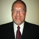 Martin, with glasses and a goatee, smiles at the camera in his dark suit, white shirt, and red patterned tie. The backdrop is a plain, light-colored wall that complements his polished look.