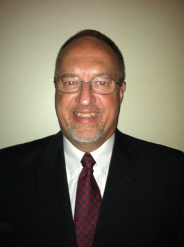 Martin, with glasses and a goatee, smiles at the camera in his dark suit, white shirt, and red patterned tie. The backdrop is a plain, light-colored wall that complements his polished look.