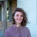 Grace, with her curly hair catching the sunlight, smiles as she stands outside a house adorned with striped brick and siding. Her red and white striped shirt mirrors the charming pattern of the home, creating a harmonious scene full of warmth and grace.