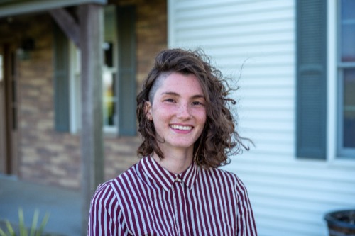 Grace, with her curly hair catching the sunlight, smiles as she stands outside a house adorned with striped brick and siding. Her red and white striped shirt mirrors the charming pattern of the home, creating a harmonious scene full of warmth and grace.