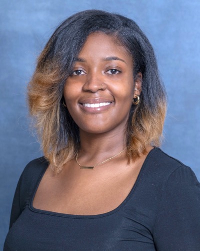 Jameala, with shoulder-length hair and a black top, smiles warmly against a blue background.