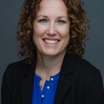 Rachel, with her curly hair, smiles at the camera in a blue blouse and black blazer. The plain gray background lends a professional appearance.