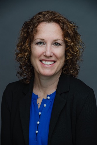 Rachel, with her curly hair, smiles at the camera in a blue blouse and black blazer. The plain gray background lends a professional appearance.