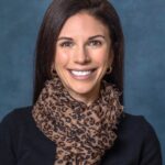 Julie, a smiling woman with long dark hair, dons a leopard-print scarf, black top, and gold hoop earrings against a blue backdrop.
