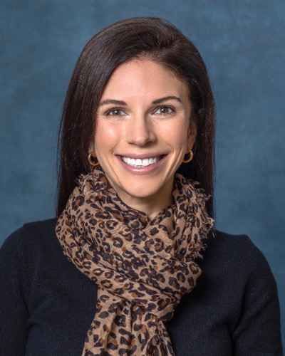 Julie, a smiling woman with long dark hair, dons a leopard-print scarf, black top, and gold hoop earrings against a blue backdrop.