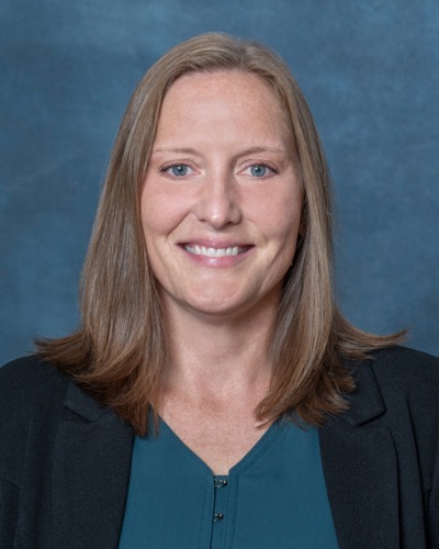 Julie Matthews, with shoulder-length brown hair and blue eyes, radiates a warm smile in a dark blazer over a teal blouse against the deep blue background.