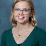 A person with shoulder-length blonde hair, glasses, and a green top smiles warmly at the camera against a calming blue-gray background. Im sorry the description lacks context for SEO keywords, but it captures a serene moment beautifully.