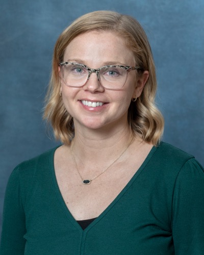 A person with shoulder-length blonde hair, glasses, and a green top smiles warmly at the camera against a calming blue-gray background. Im sorry the description lacks context for SEO keywords, but it captures a serene moment beautifully.