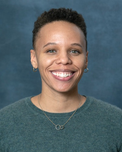 A smiling person with short hair, clad in a green sweater and a circular pendant necklace, exudes charm against the blue background. This delightful moment captures Chloes radiant presence.