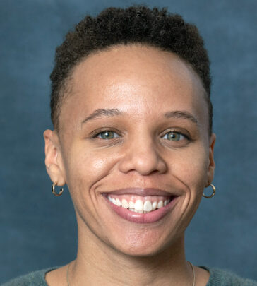 Chloe, a smiling person with short curly hair and small hoop earrings, poses against a blue-gray background.