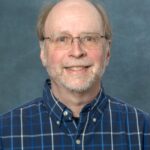 A smiling man with glasses, a beard, and receding hair wears a blue plaid shirt against a plain gray background, capturing the essence of Jeffreys easygoing style.