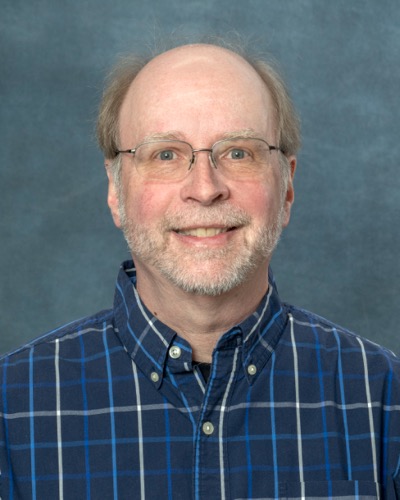A smiling man with glasses, a beard, and receding hair wears a blue plaid shirt against a plain gray background, capturing the essence of Jeffreys easygoing style.