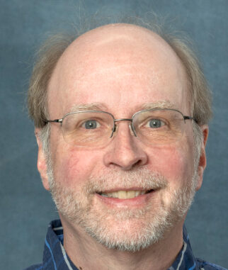 A smiling man with glasses, a light beard, and short hair stands against a gray-blue background. He is wearing a blue checkered shirt. Im sorry for any inconvenience, but I would be happy to help if you could provide more details or context for SEO optimization.