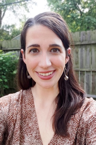 Maria, a woman with long dark hair, is smiling at the camera. Shes wearing a brown patterned top and drop earrings. The backdrop of a wooden fence and lush greenery hints at an outdoor setting, creating a scene Paschke might capture beautifully.