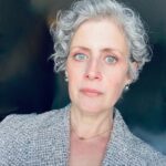A person with short gray hair and blue eyes, perhaps named Preston or Rebecca, looks directly at the camera. They wear a textured gray jacket and small earrings, standing against a dark background.